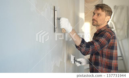 Male construction worker wearing protective gloves smoothing plaster onto wall surface during home renovation using professional trowel tool 137731944