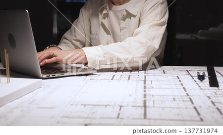 Architect or engineer working late, typing on laptop, reviewing blueprints and technical drawings for a new construction project or building plan in an office Architect or engineer working late, typing on laptop, reviewing blueprints and technical drawings for a new construction project or building plan in an office 137731974