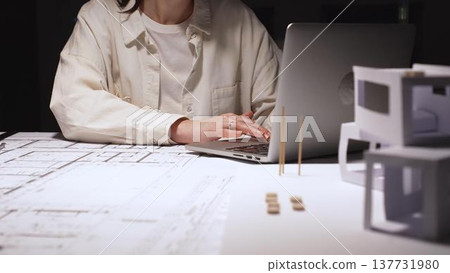 Architect or engineer working late, typing on laptop, reviewing blueprints and technical drawings for a new construction project or building plan in an office Architect or engineer working late, typing on laptop, reviewing blueprints and technical drawings for a new construction project or building plan in an office 137731980