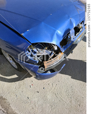 A wrecked car rests on the street with a damaged front end. The paint is chipped and rust is showing. The area looks deserted with no people around. 137732385