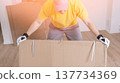 A worker unpacks furniture from cardboard boxes. Man lifts large cardboard box with effort. Hands grip sides, wearing white gloves. Bright yellow shirt contrasts with soft lighting. Gentle smile 137734369