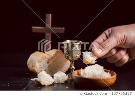 Communion scene with hand offering bread beside ornate chalice and wooden cross on dark background, symbolizing Christian Eucharist, worship, faith, sacrifice and remembrance with copy space. Communion scene with hand offering bread beside ornate chalice and wooden cross on dark background, symbolizing Christian Eucharist, worship, faith, sacrifice and remembrance with copy space. 137735397