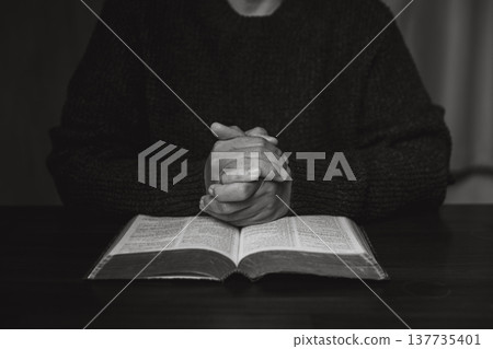 Black and white close-up of praying hands above an open Bible with a wooden cross on a table, symbolizing Christian faith, devotion, worship and spiritual reflection in a quiet home setting. 137735401