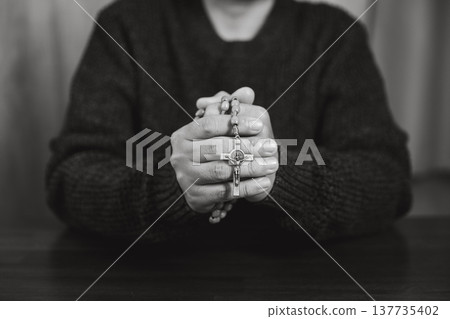 Black and white close-up of hands holding rosary beads with cross, symbolizing prayer, worship, Catholic faith, devotion and quiet spiritual reflection. Soft background with copy space for text. 137735402