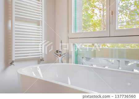 A modern bathroom featuring a white bathtub, a towel radiator, and large windows with a view of greenery outside. A modern bathroom featuring a white bathtub, a towel radiator, and large windows with a view of greenery outside. 137736560