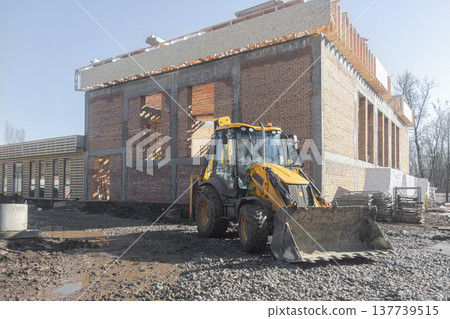 A backhoe loader works at a construction site next to an unfinished brick building with a wood-frame roof A backhoe loader works at a construction site next to an unfinished brick building with a wood-frame roof 137739515