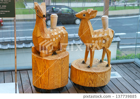 Nara, Japan - Sep 26 2024, close-up view of sculptures carved from wood, two deer sitting and standing on tree stumps and coffee cups on top of their backs, with driving car on background, Nara, Japan Nara, Japan - Sep 26 2024, close-up view of sculptures carved from wood, two deer sitting and standing on tree stumps and coffee cups on top of their backs, with driving car on background, Nara, Japan 137739885