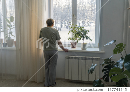 Elderly woman standing by a window, looking out at the snow-covered trees during a cold winter day, feeling solitude 137741345