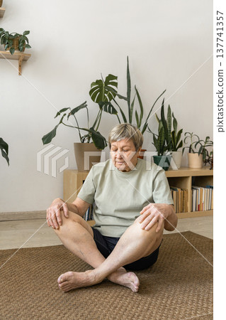 Senior woman sitting in a mindful lotus pose on a yoga mat, practicing meditation and relaxation in her living room. Mindful happiness and active living for seniors 137741357