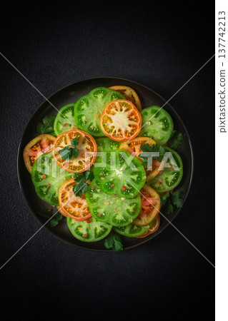 Sliced green tomatoes, on a dark surface, garnished with herbs and salt, top view, Sliced green tomatoes, on a dark surface, garnished with herbs and salt, top view, 137742213