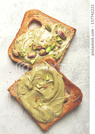 Two slices of toasted bread, topped with pistachio paste and sprinkled with pistachios, spread on a light surface Two slices of toasted bread, topped with pistachio paste and sprinkled with pistachios, spread on a light surface 137742222
