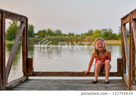 Young girl smiling on a wooden dock by a calm lake during golden hour Young girl smiling on a wooden dock by a calm lake during golden hour 137742614