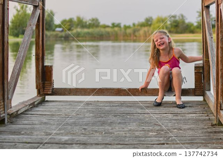 Joyful young girl laughing on a wooden pier by the tranquil lake during golden hour. A happy child with blonde hair sits on a weathered wooden dock, her face lit up with laughter, with a calm lake and Joyful young girl laughing on a wooden pier by the tranquil lake during golden hour. A happy child with blonde hair sits on a weathered wooden dock, her face lit up with laughter, with a calm lake and 137742734