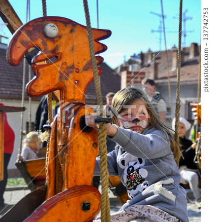 Young girl with face paint enjoys a ride on a colorful wooden swing at a festival Young girl with face paint enjoys a ride on a colorful wooden swing at a festival 137742753