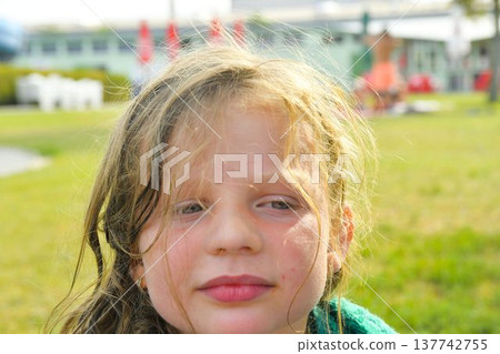 Close-up portrait of a young girl with windswept blonde hair outdoors on a sunny day. A young girl with tousled blonde hair and rosy cheeks looks towards the camera with a gentle expression, enjoying 137742755