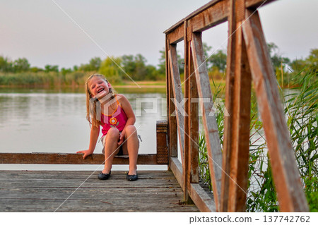 Joyful little girl laughing on a wooden pier by the lake at sunset 137742762