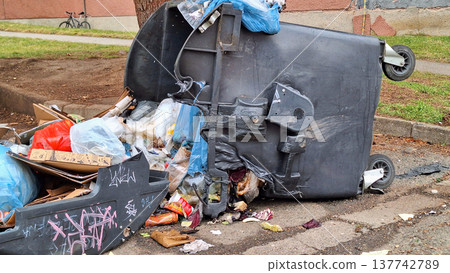Overturned garbage bin spilling trash onto the street, waste management issues 137742789