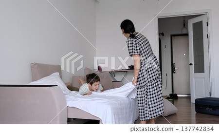 A mother laughs as she lifts a white sheet over her son on a wide bed, captured candidly with soft daylight. The moment celebrates routines that build comfort, bonding, and resilience in a real home. A mother laughs as she lifts a white sheet over her son on a wide bed, captured candidly with soft daylight. The moment celebrates routines that build comfort, bonding, and resilience in a real home. 137742834