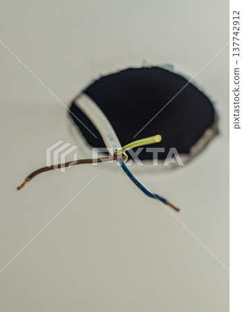 Exposed electrical wires hanging from a round hole in a white ceiling for lighting installation. Exposed electrical wires hanging from a round hole in a white ceiling for lighting installation. 137742912