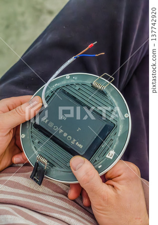 Electrician holding the back side of a round LED recessed light fixture before ceiling installation. 137742920