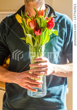 Man holding a glass vase full of vibrant red and yellow spring tulips in bright natural sunlight. 137742921