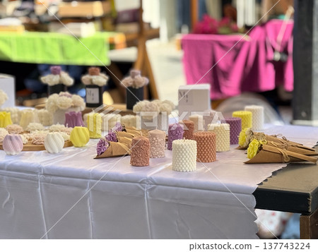 Pastel handmade candles and floral bouquets are arranged on a market table outdoors. Handcrafted decor, softness, gift culture, spring mood, and the delicate appeal of artisanal design. Pastel handmade candles and floral bouquets are arranged on a market table outdoors. Handcrafted decor, softness, gift culture, spring mood, and the delicate appeal of artisanal design. 137743224