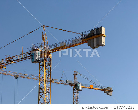 Tower cranes rise above a construction site under a clear blue sky. Engineering, development, urban expansion, industrial progress, structural planning, and the geometry of modern building activity. Tower cranes rise above a construction site under a clear blue sky. Engineering, development, urban expansion, industrial progress, structural planning, and the geometry of modern building activity. 137743233