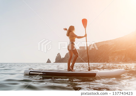 Woman Paddleboarding Ocean Coastline - A woman paddleboards in the ocean with a scenic coastline in the background. Woman Paddleboarding Ocean Coastline - A woman paddleboards in the ocean with a scenic coastline in the background. 137743423