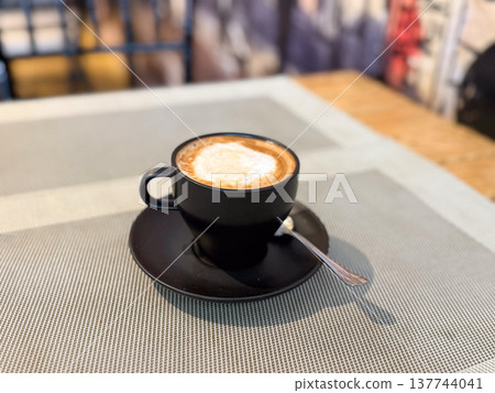 Black cup of cappuccino on a table in a quiet cafe interior. Morning pause, warmth, caffeine ritual, minimal presentation, and the calm pleasure of a simple coffee moment. Black cup of cappuccino on a table in a quiet cafe interior. Morning pause, warmth, caffeine ritual, minimal presentation, and the calm pleasure of a simple coffee moment. 137744041