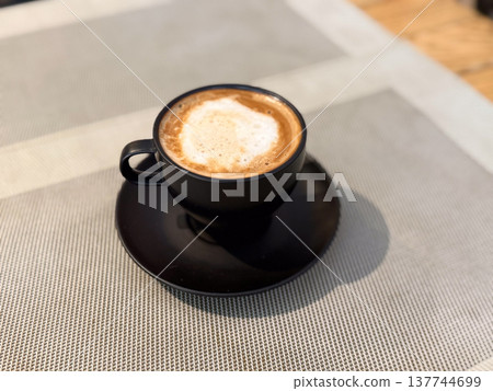 Black cup of cappuccino on a table in a quiet cafe interior close-up. Morning pause, warmth, caffeine ritual, minimal presentation, and the calm pleasure of a simple coffee moment. 137744699