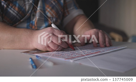 Hands are writing letters on a paper using a red marker at a table. There are blue pens and blank paper around in dim lighting. The person is focused on the task. Hands are writing letters on a paper using a red marker at a table. There are blue pens and blank paper around in dim lighting. The person is focused on the task. 137745009