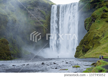 Huge waterfall surrounded by mossy rocky cliff and people walking towards it, Iceland Huge waterfall surrounded by mossy rocky cliff and people walking towards it, Iceland 137745052