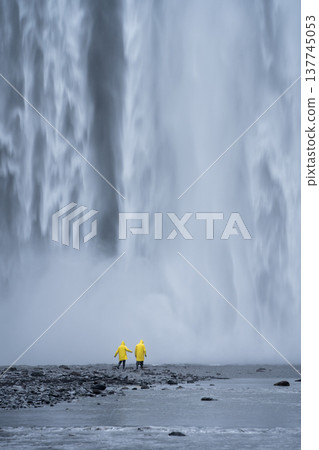 Vertical shot of couple in yellow raincoats walking in front of huge waterfall, Iceland 137745053