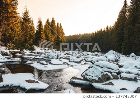 A serene winter evening showcases the Jizera River along the Czechia and Poland border. Snow blankets the rocks and trees while the water flows gently, creating a peaceful scene. 137745307