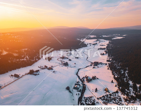 Aerial view shows Jizerka mountain village covered in snow. The landscape features houses, trees, and winding paths under a bright sky at sunset. 137745312