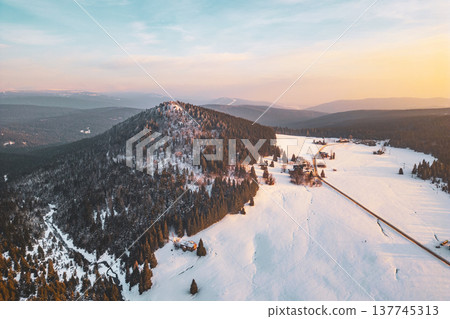 Drone captures Bukovec Mountain in Jizera Mountains covered in snow. The landscape includes dense forests and an open snowy field. The sun sets in the background, creating a warm glow. 137745313