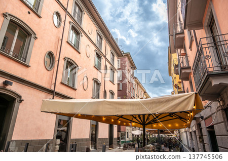 Milan, italy, april 1, 2024. Brera street cafe under a large umbrella amid pastel historic buildings, cobblestone alley evoking charming milanese city life Milan, italy, april 1, 2024. Brera street cafe under a large umbrella amid pastel historic buildings, cobblestone alley evoking charming milanese city life 137745506