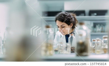 Female scientist analyzing samples in laboratory workspace, focused research scene with glass bottles and equipment representing chemistry investigation, biotechnology and medical science 137745814