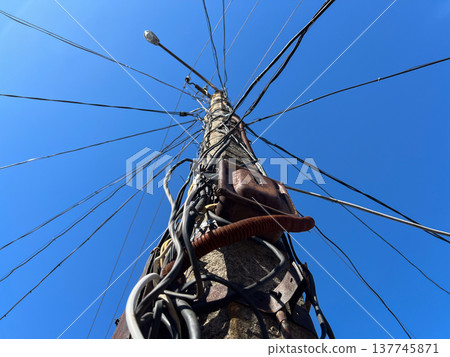 Beneath a vast blue sky, a sturdy concrete pole stands tall with numerous wires sprawling outwards, connecting homes and powering the community Beneath a vast blue sky, a sturdy concrete pole stands tall with numerous wires sprawling outwards, connecting homes and powering the community 137745871