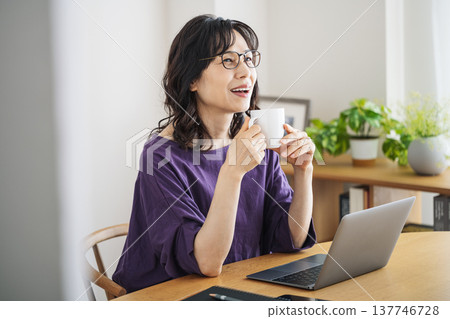 一位五十多歲的中年婦女在家裡的客廳裡打開筆記型電腦,一邊喝咖啡。 一位五十多歲的中年婦女在家裡的客廳裡打開筆記型電腦,一邊喝咖啡。 137746728