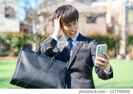 一位年輕的商人，看著他的智慧型手機，痛苦地抱住頭（上半身）。 137746800