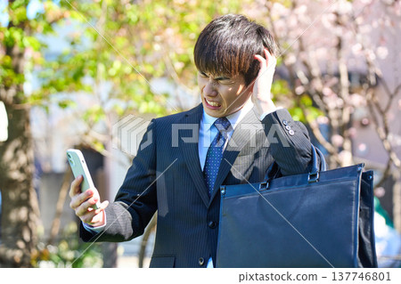 一位年輕的商人神情沮喪，雙手抱頭，低頭看著手機，他身處櫻花盛開的公園。 （上半身） 137746801