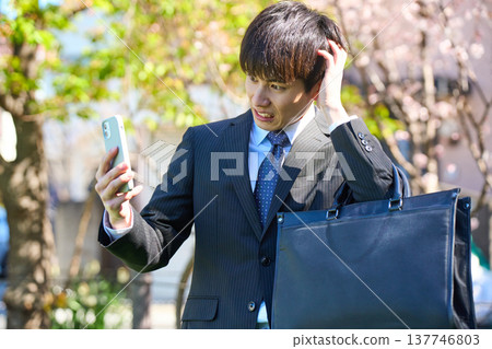 A young businessman, looking dejected and holding his head in his hands while looking at his smartphone, in a park with cherry blossoms in bloom. (Upper body) A young businessman, looking dejected and holding his head in his hands while looking at his smartphone, in a park with cherry blossoms in bloom. (Upper body) 137746803