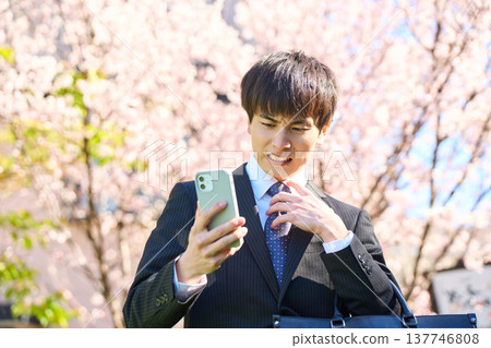 一位年輕的商人，在櫻花盛開的公園裡，低頭看著手機，一臉苦惱。 （上半身） 137746808