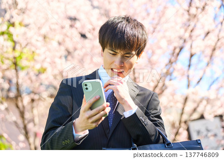 一位年輕的商人，在櫻花盛開的公園裡，低頭看著手機，一臉苦惱。 （上半身） 137746809