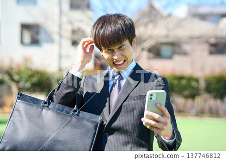 A young businessman, looking at his smartphone and holding his head in distress (upper body). 137746812