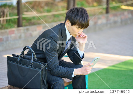 A young businessman, looking dejected while looking at his smartphone on a park bench (upper body). 137746945