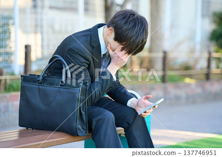 A young businessman, looking dejected while looking at his smartphone on a park bench (upper body). A young businessman, looking dejected while looking at his smartphone on a park bench (upper body). 137746951