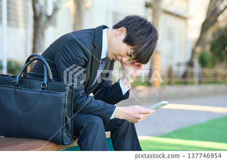 A young businessman, looking dejected while looking at his smartphone on a park bench (upper body). A young businessman, looking dejected while looking at his smartphone on a park bench (upper body). 137746954