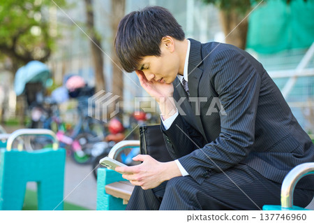 一位年輕的商人，神情沮喪地坐在公園長椅上看著他的智慧型手機（上半身）。 137746960
